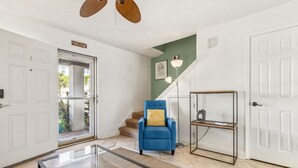 Amplia sala de estar con una mesa de centro de vidrio, un sillón azul con cojín amarillo, una consola de metal y una escalera con un muro decorado en tonos verdes.