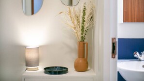 Une salle de bain avec un miroir, un vase contenant des plantes séchées et une lampe posée sur une étagère.