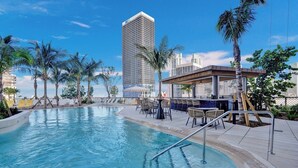 Una piscina con sillas de descanso y una zona de bar, rodeada de palmeras y edificios modernos.