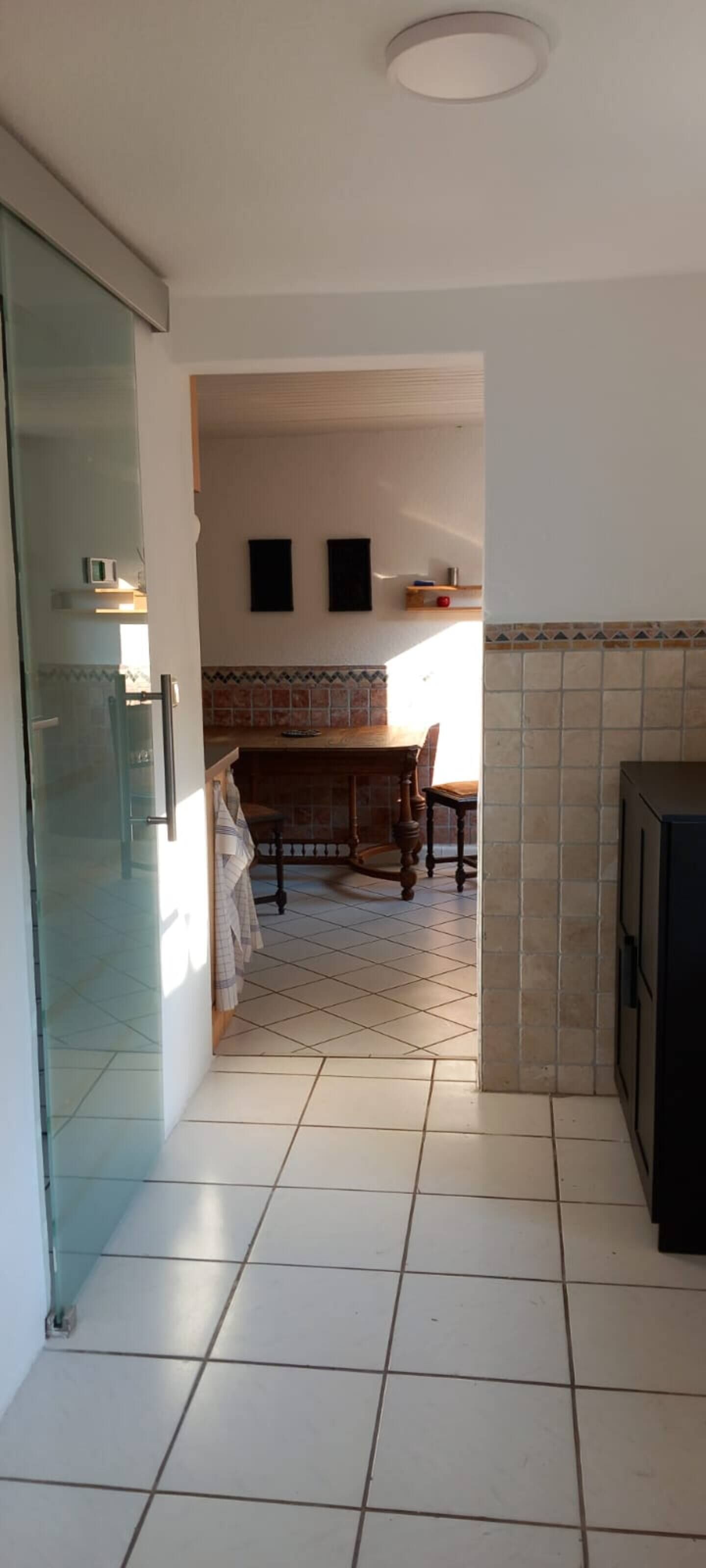 Ein schmaler Gang führt zu einem Essbereich mit Fliesenboden, einem Holztisch und Stühlen. Auf der linken Seite befindet sich eine Glastür, und auf der rechten eine schwarze Schrank.