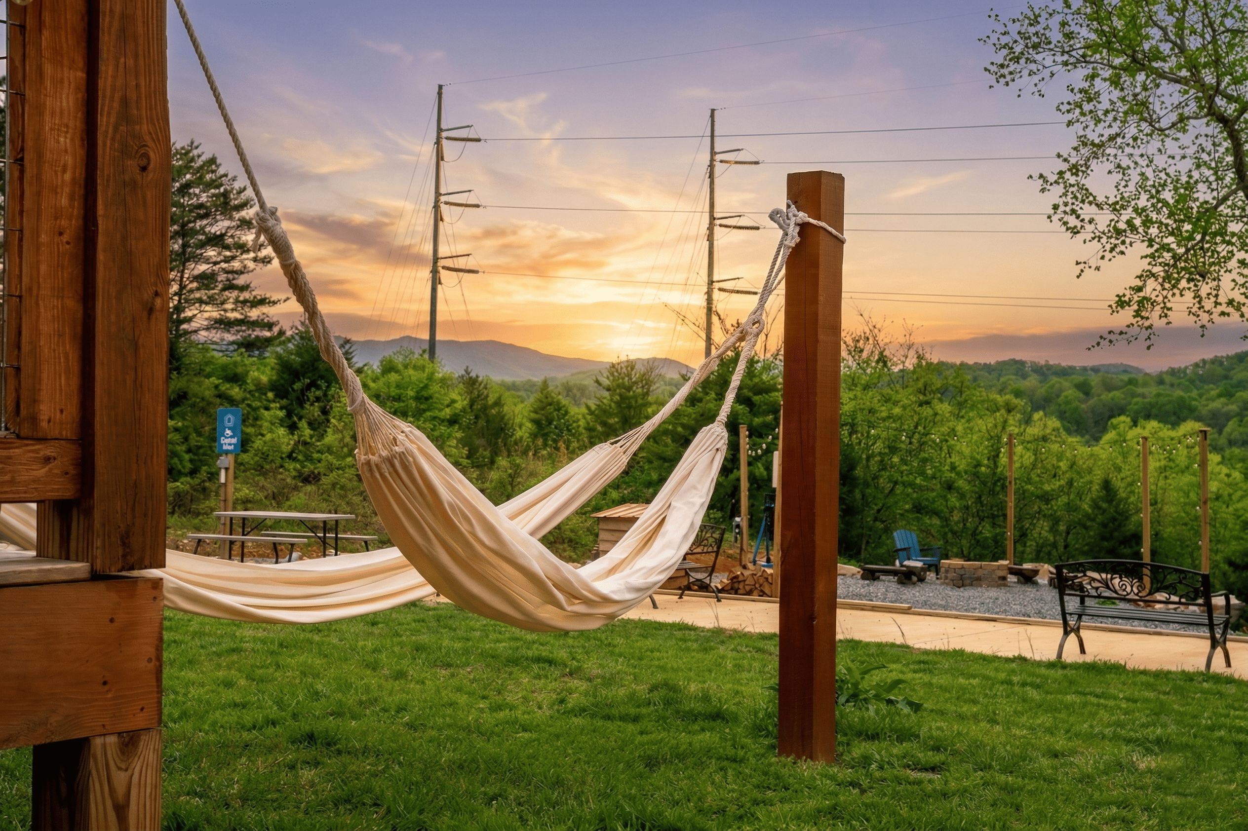 A hammock tied to a wooden post in a grassy area with a picnic table and benches, set against a backdrop of trees and a sunset.