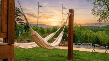 A hammock tied to a wooden post in a grassy area with a picnic table and benches, set against a backdrop of trees and a sunset.