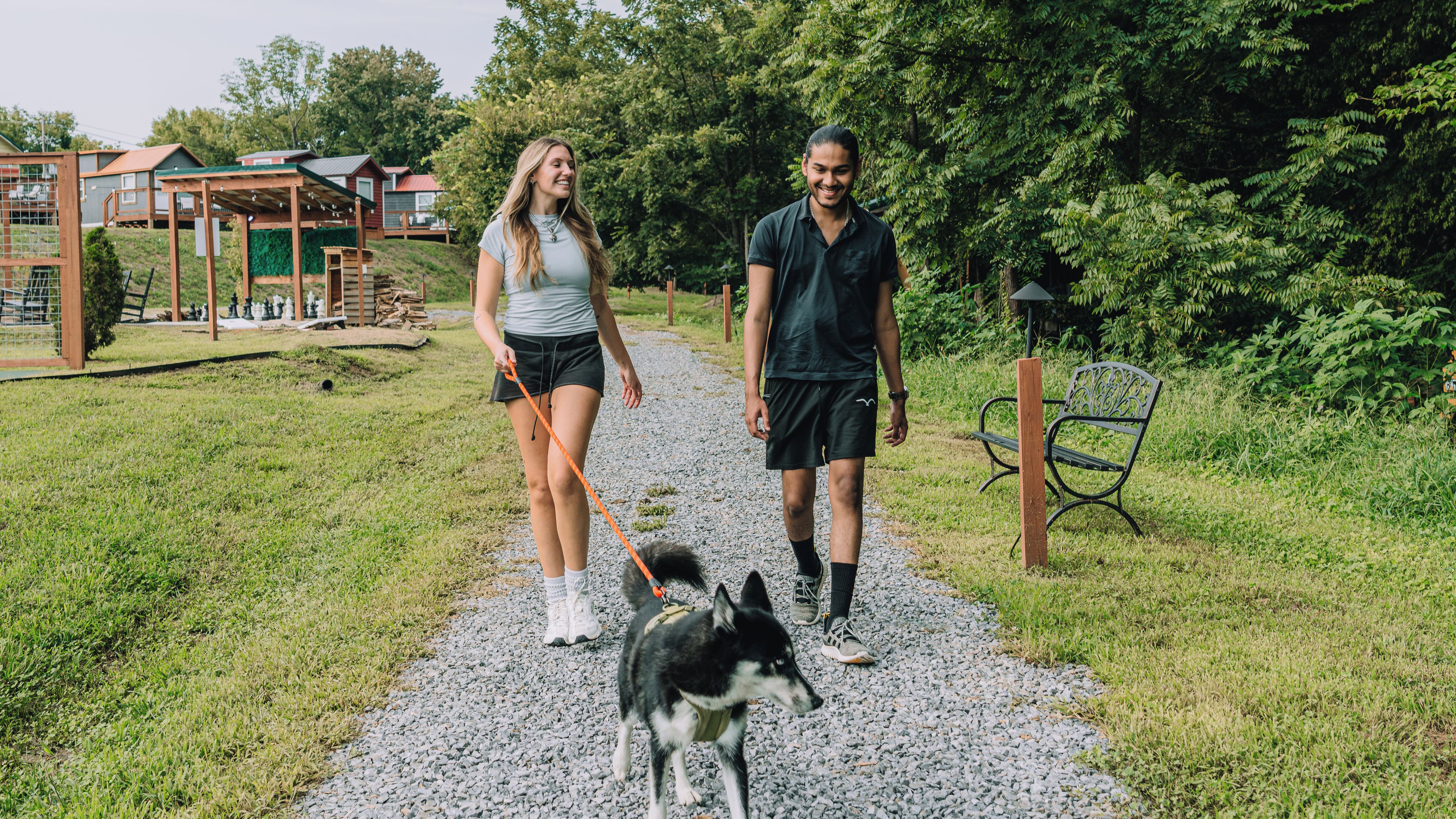 Deux personnes promènent un chien sur un chemin de gravier, avec en arrière-plan de la verdure et des structures en bois.