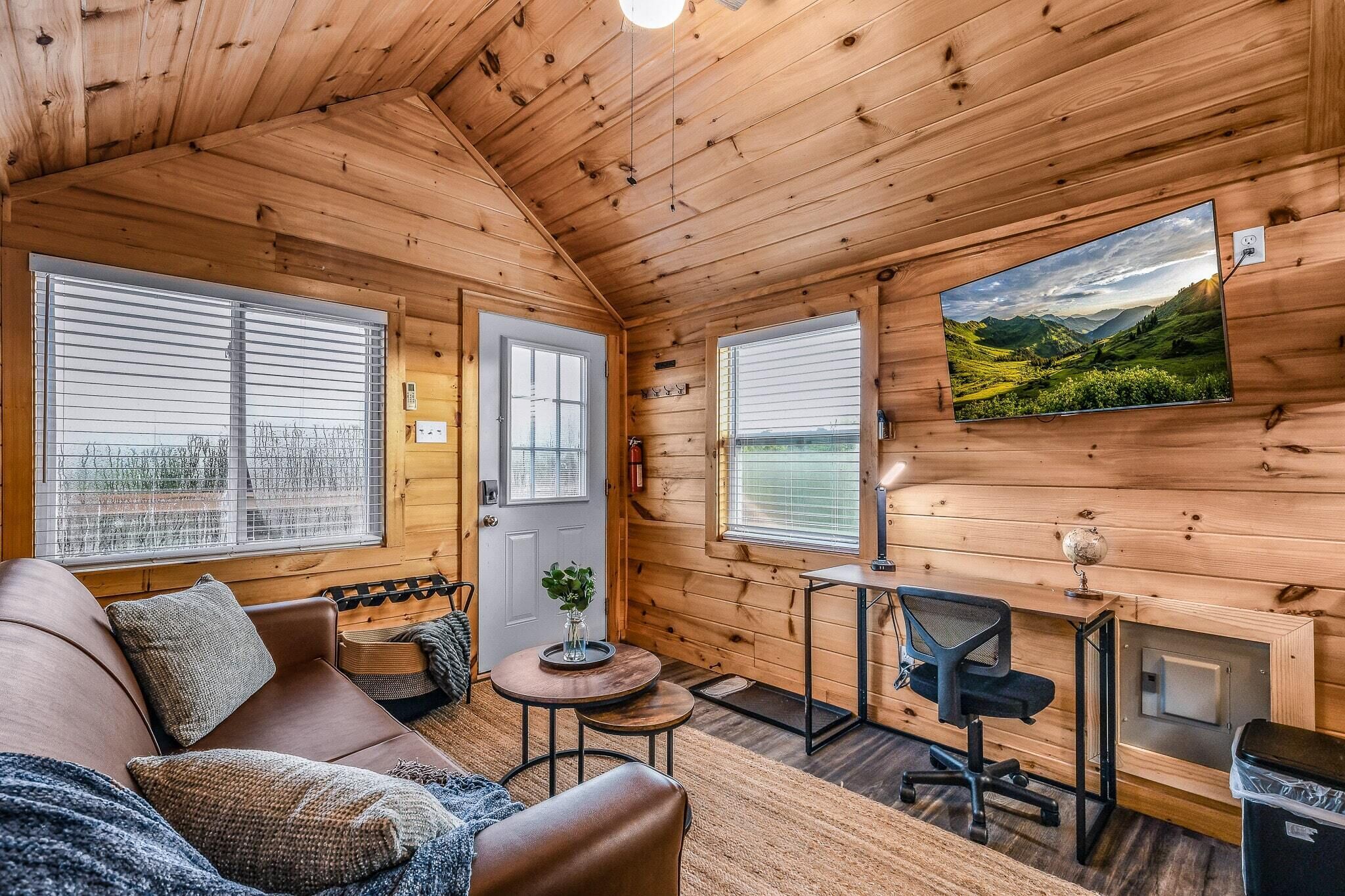 A cozy cabin interior with a wooden ceiling, a sofa, a small round table, a desk with a chair, and a large window with a scenic view.