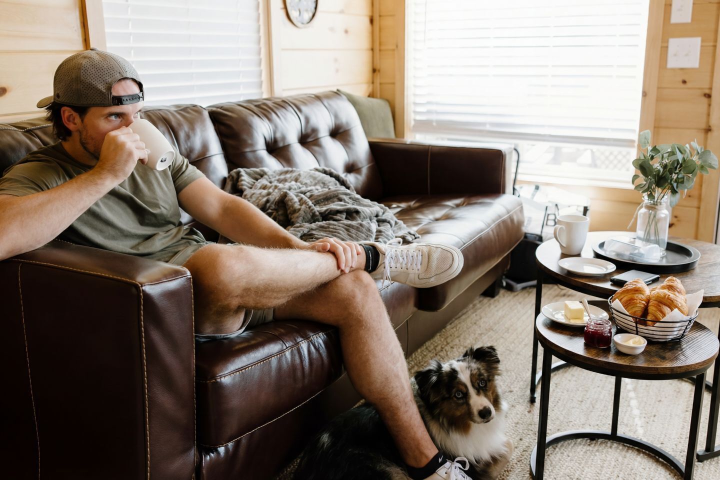 A person sitting on a leather couch with a dog, a table with croissants, and a small vase with plants.