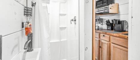 Une salle de bain avec douche, lavabo et un coin cuisine doté de meubles et d’appareils électroménagers en bois.