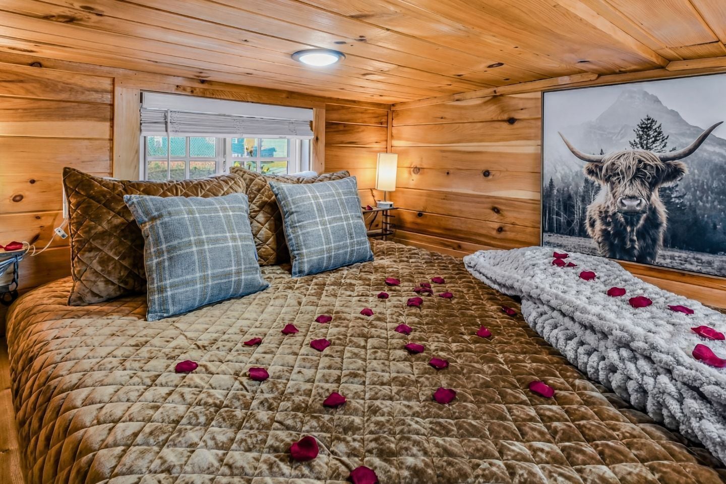 A cozy wooden cabin bedroom with a large bed, a nightstand, a lamp, and a painting of a mountain and bull.