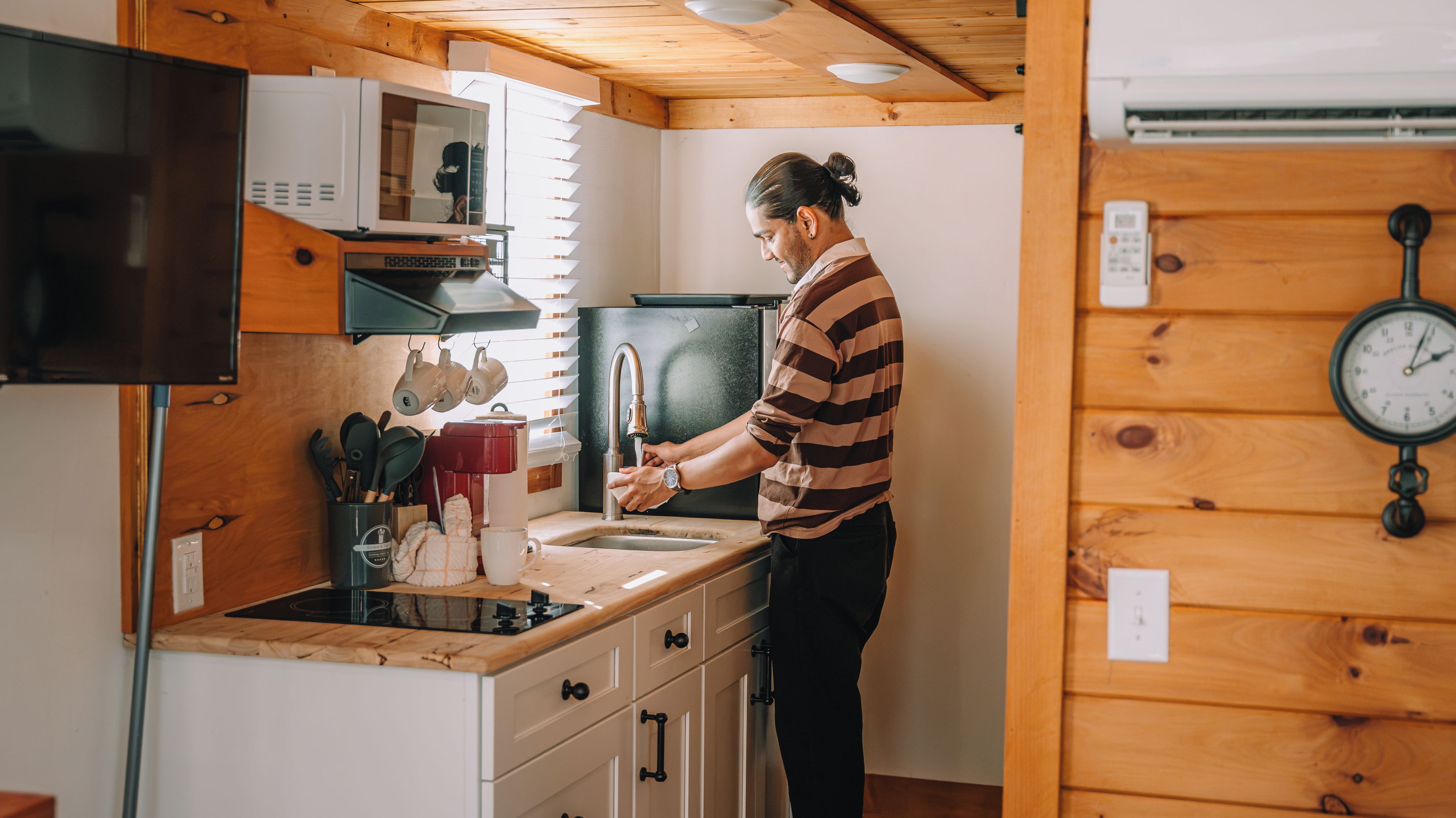 Une personne est en train de faire la vaisselle dans une cuisine aux murs en bois, où l’on peut voir une horloge et un four à micro-ondes.