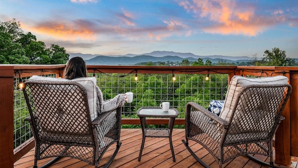 A wooden deck with wicker rocking chairs and a small table, overlooking a scenic landscape with mountains and trees.