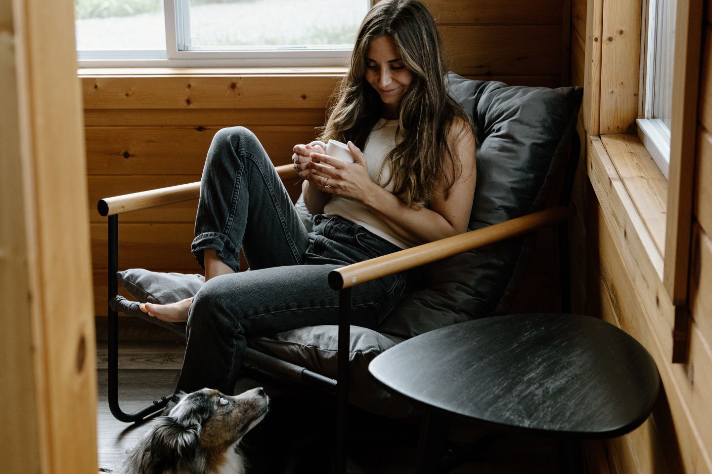 A person sitting on a chair with a dog beside them, in a room with wooden walls and a window.