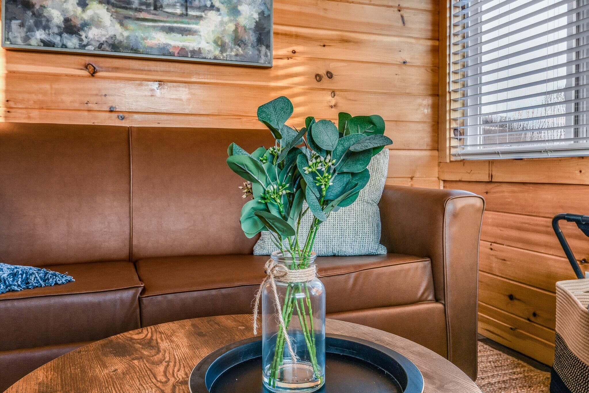 Un intérieur en bois avec un canapé en cuir marron, un vase en verre transparent contenant des plantes vertes et un tableau accroché au mur.