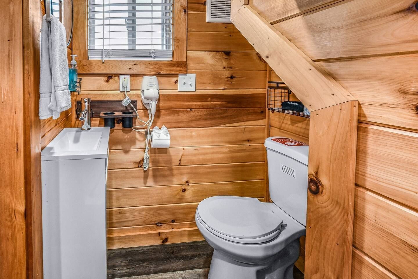 Een klein houten badkamertje met een wastafel, toilet en een raam boven de wastafel.