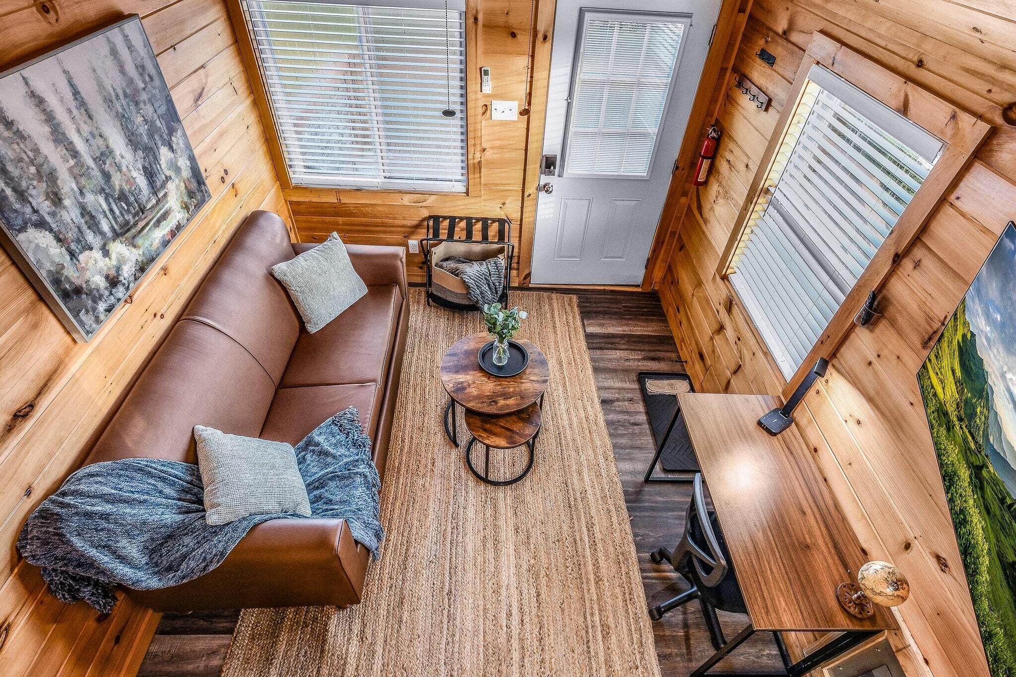 Interior acogedor de una cabaña con piso de madera, un sofá de cuero marrón con cojines, una mesita redonda con una lámpara y una puerta con ventanal.