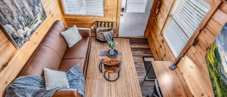 A cozy cabin interior with a wooden floor, a brown leather sofa with cushions, a small round table with a lamp, and a door with a window.