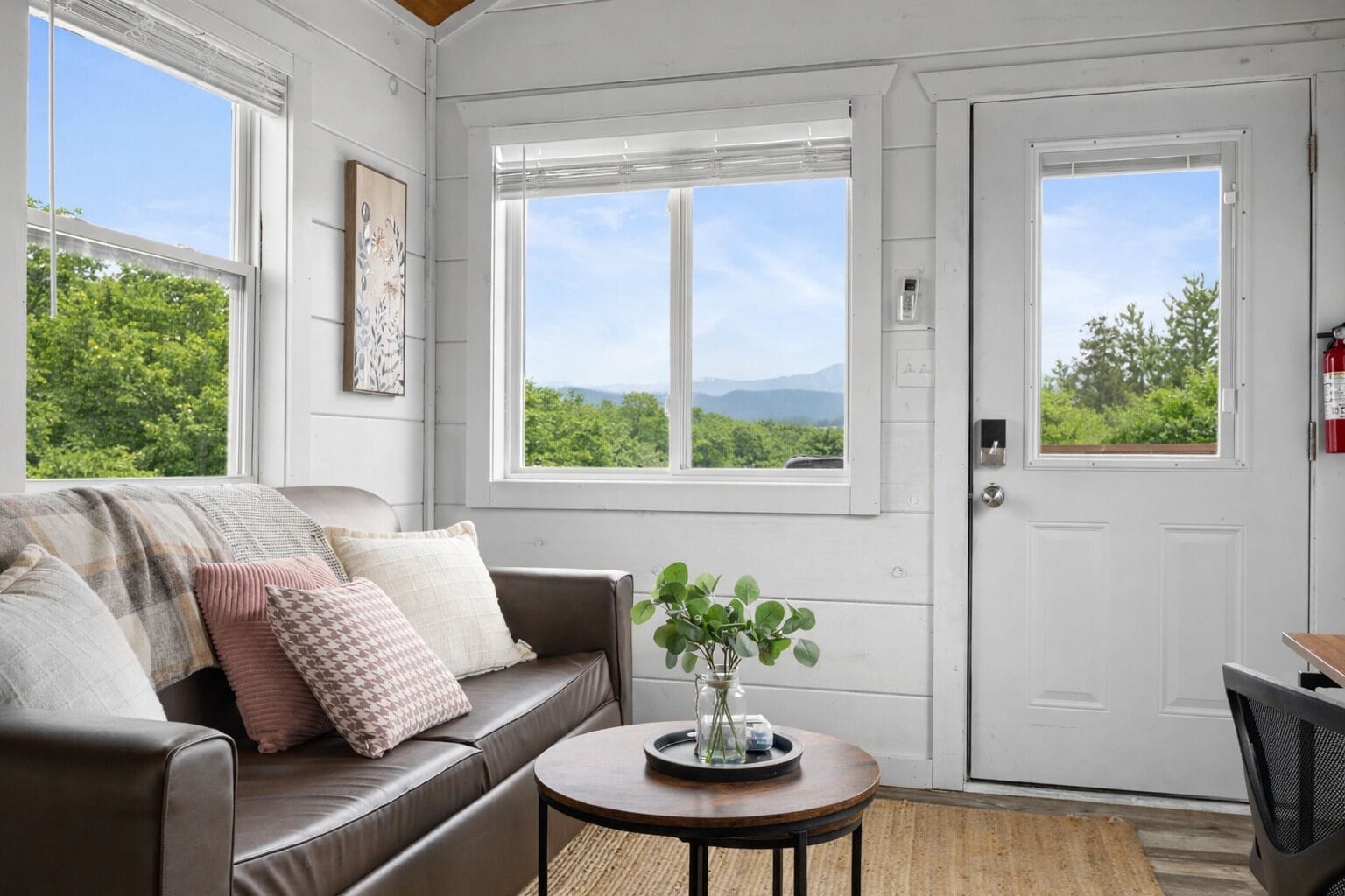 A room with a brown leather sofa, a small round table with a vase, and two windows with a view of greenery and mountains.