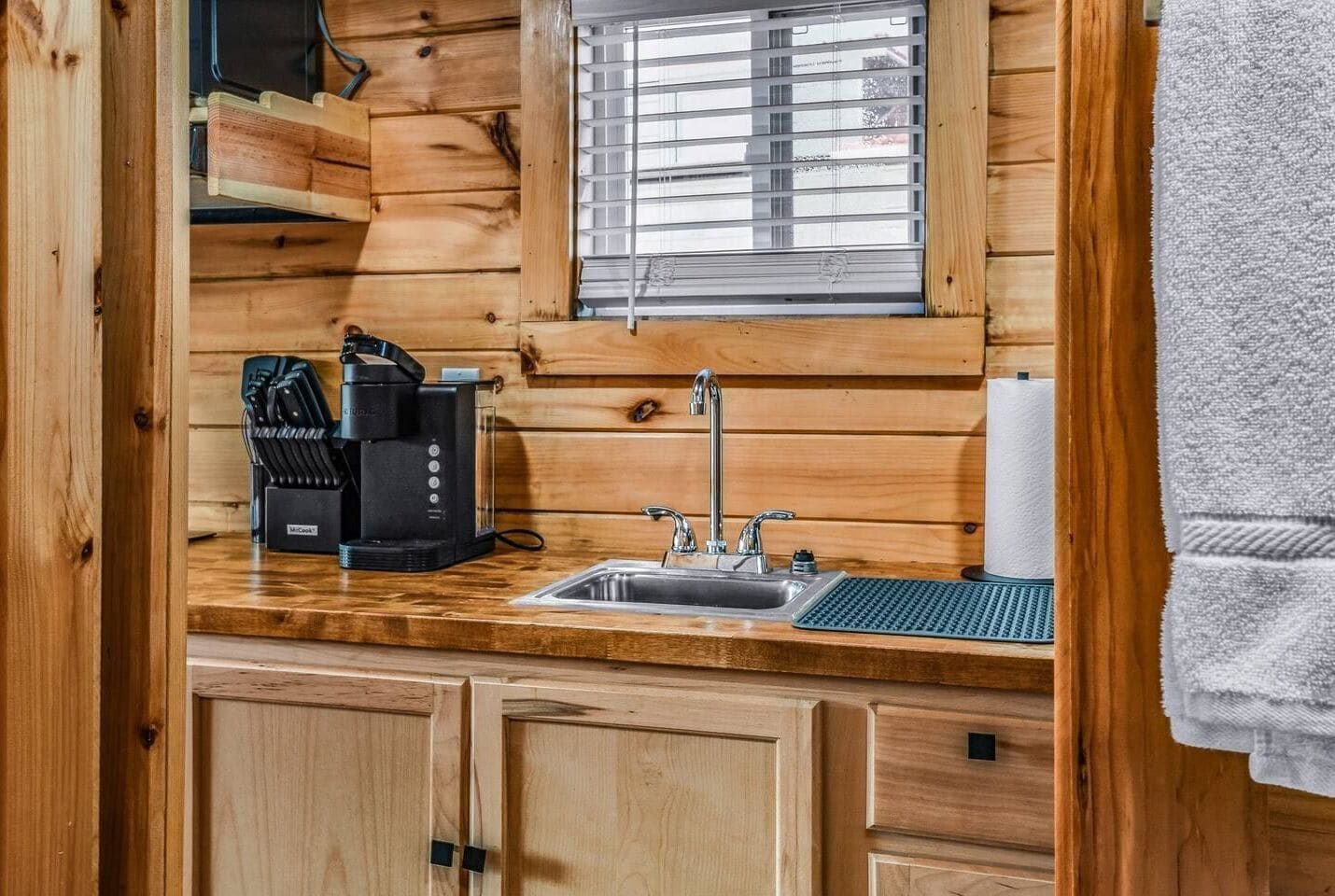 A wooden cabin-style kitchen with a built-in sink, a coffee maker, and a window with blinds.