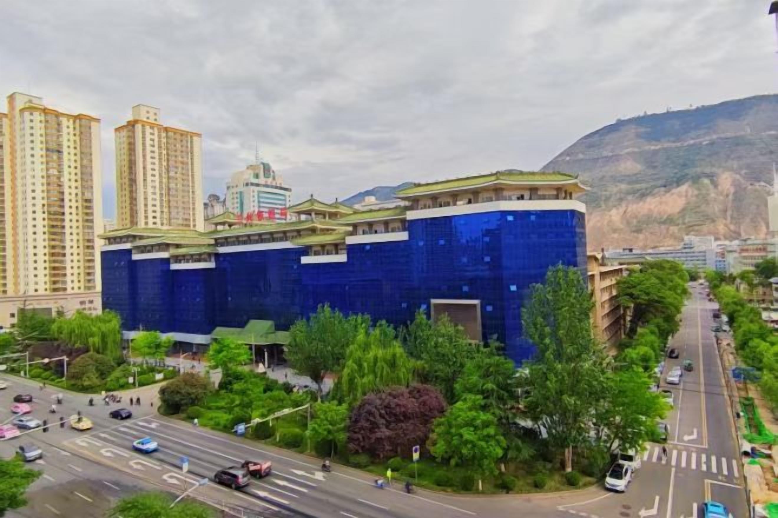 A multi-story building with a blue facade, surrounded by greenery and a busy street.