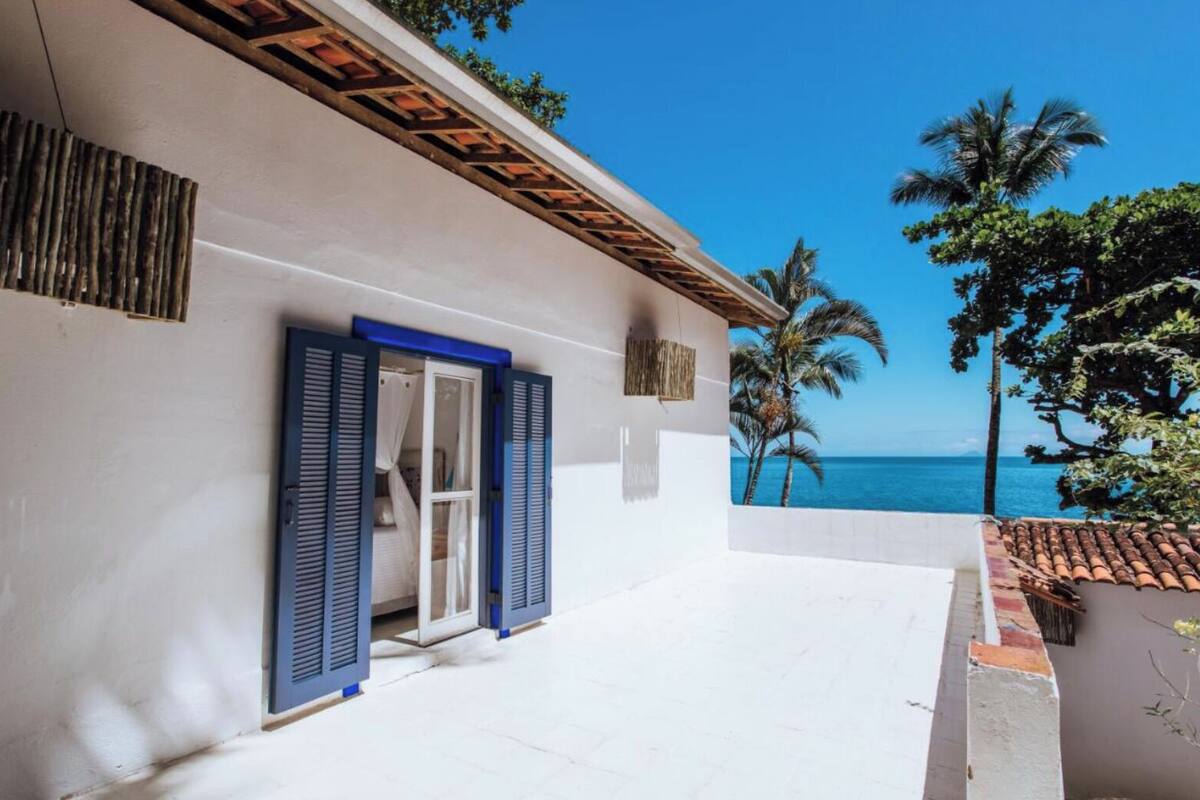 Un bâtiment situé sur la côte, doté d’un balcon donnant sur la mer, avec une façade blanche, des volets bleus et une vue sur les palmiers.