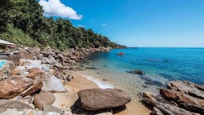 A rocky beach with clear blue water, a wooden pier, and dense greenery. - Blow Show Casa I  (Ilhabela)