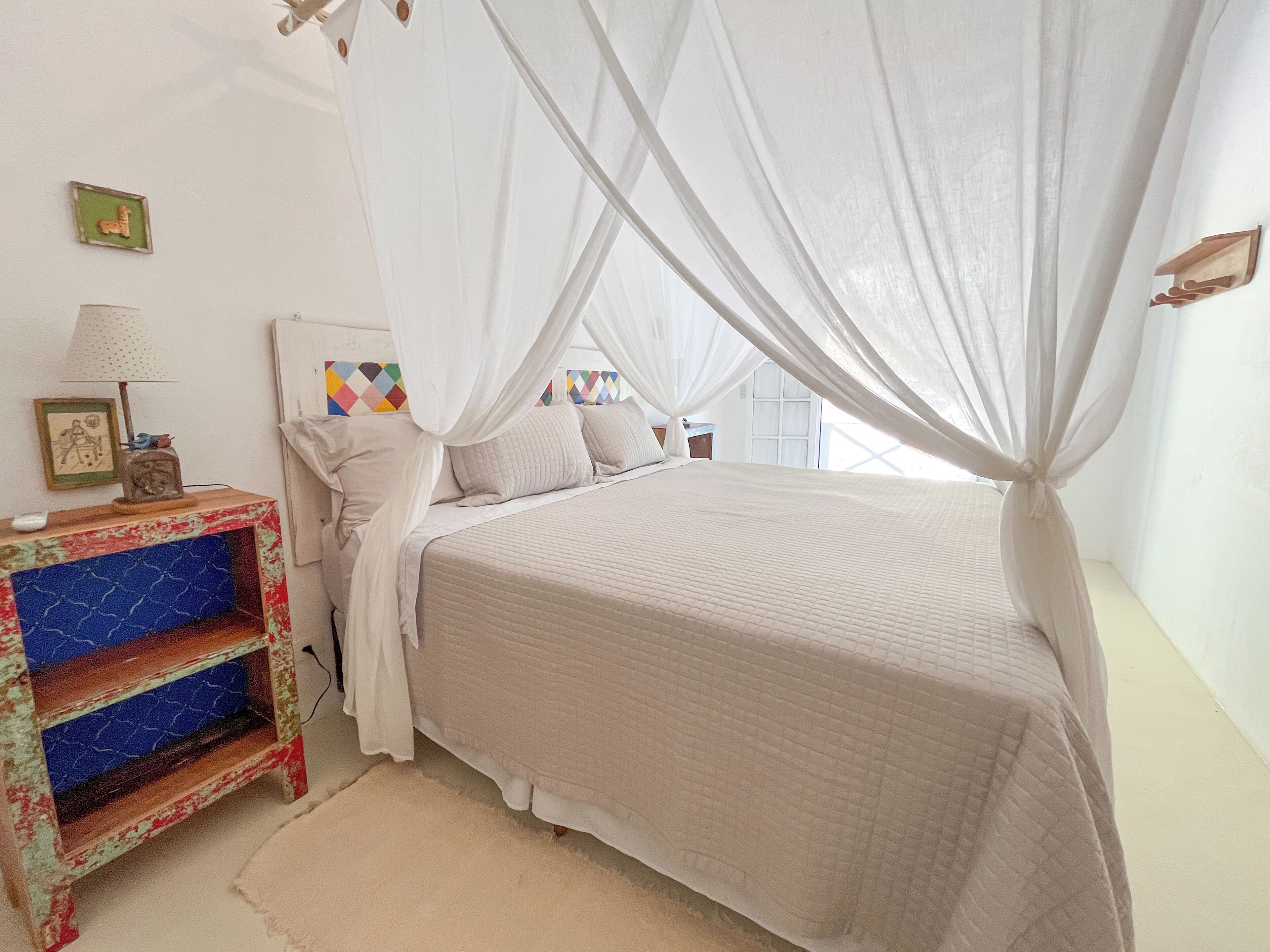 A bedroom with a canopy bed, a bedside table with a lamp, and a colorful shelf.