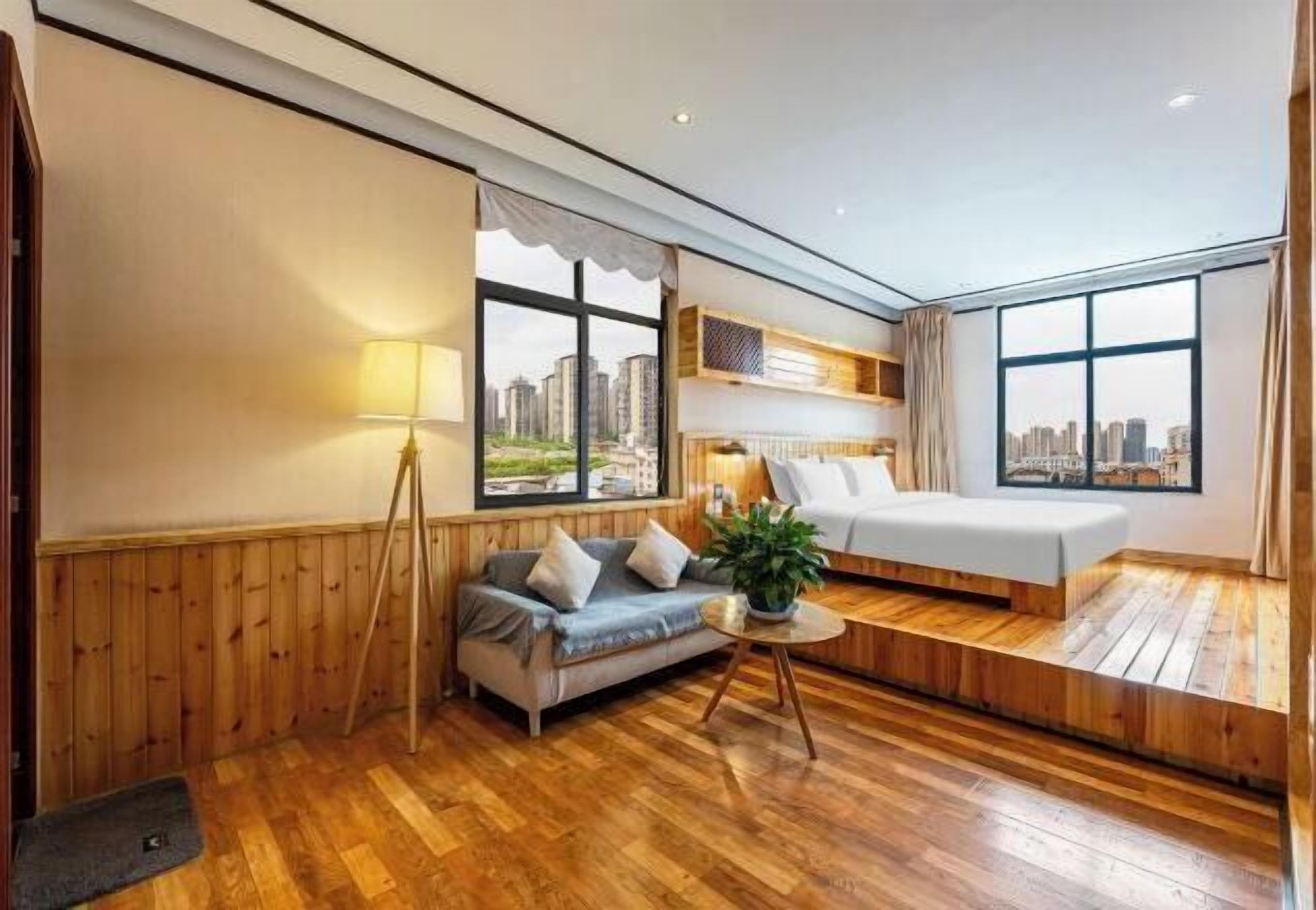 Ein modernes Hotelzimmer mit Holzboden, einer Couch, einem Bett, einem kleinen Tisch und Blick auf die Stadt durch große Fenster.