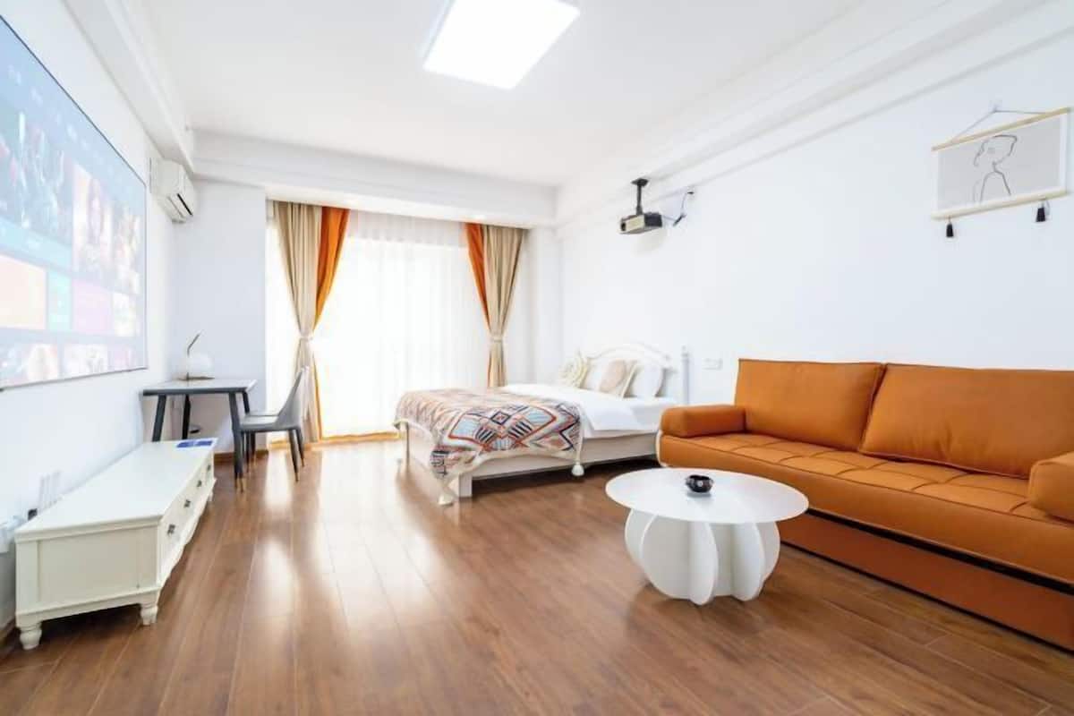 A hotel room with a bed, an orange sofa, a white coffee table, a desk, and a projector screen.