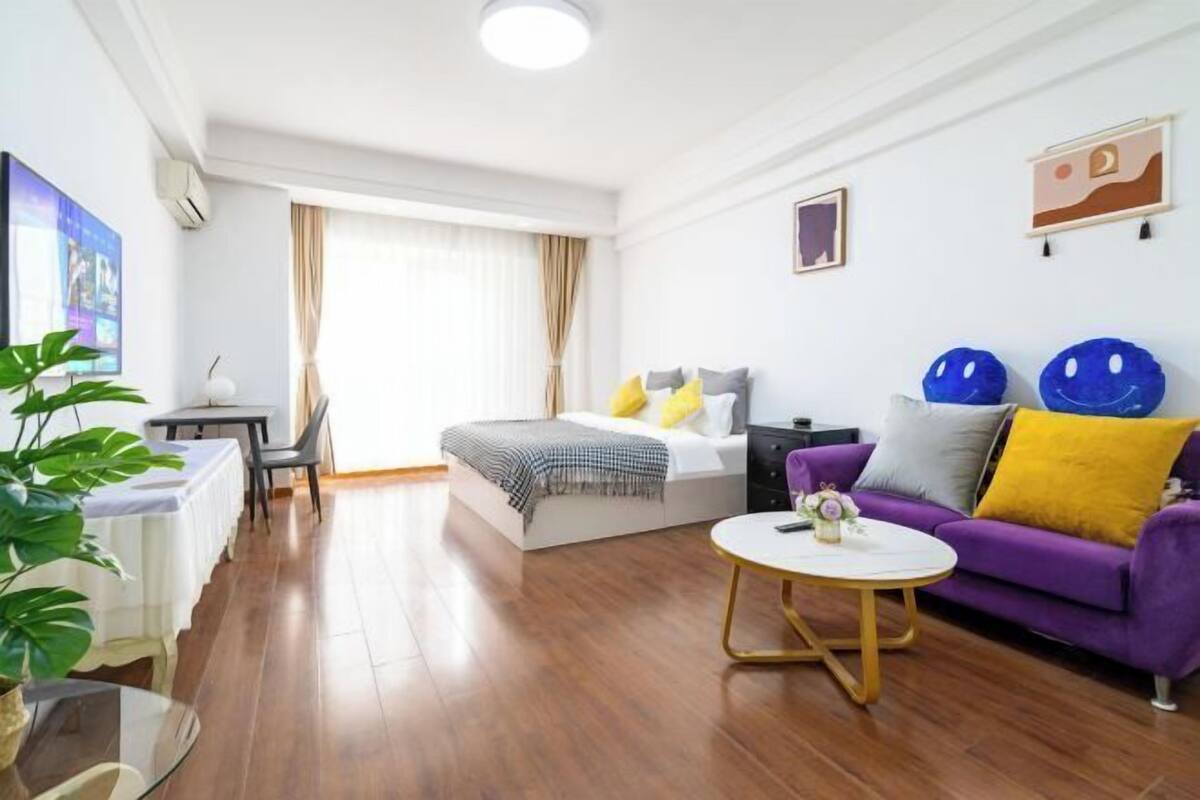 A modern hotel room with a bed, a purple sofa, a round coffee table, and a TV on the left wall.