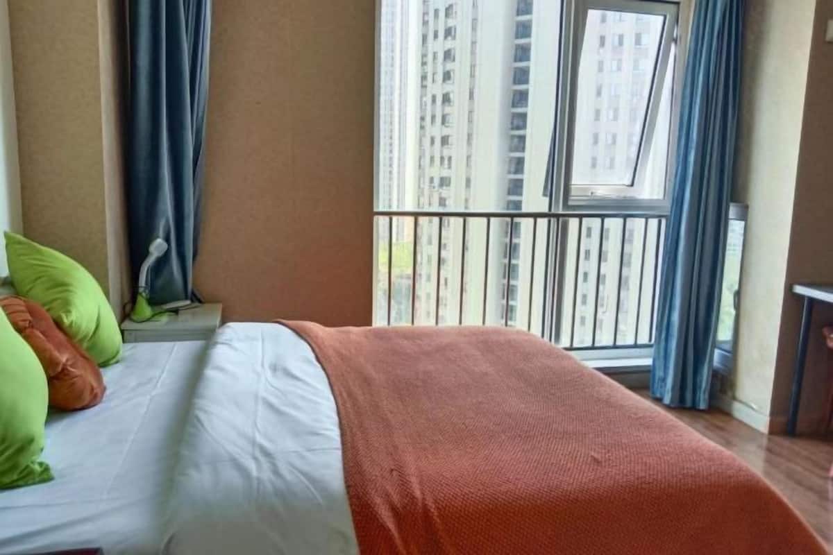 A hotel room with a bed, pillows, a window with a view of tall buildings, and a balcony.