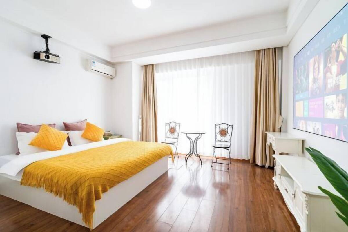 A hotel room with a bed, a TV, a projector, a small table, chairs, and a large window with curtains.