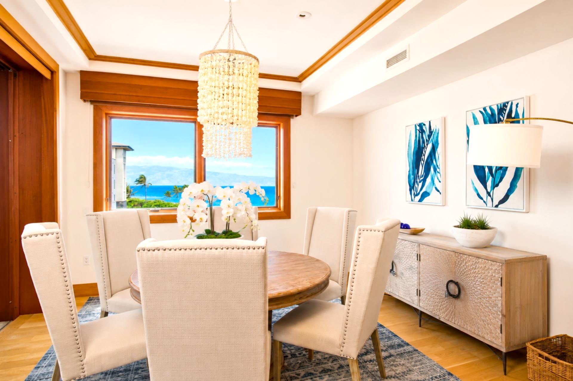 A dining area with a round table, six chairs, a chandelier, a view of the ocean, and artwork on the wall.