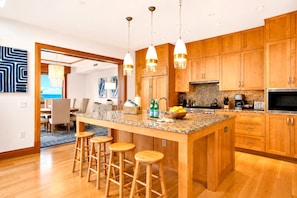 Una cocina moderna con armarios de madera, encimera de granito y electrodomésticos de acero inoxidable. Se ven tres taburetes de bar y una zona de comedor.