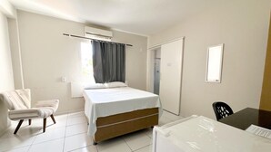 A bedroom with a bed, a chair, a desk, a refrigerator, a window with curtains, and a door. - HOTEL OITICICA (Fortaleza)
