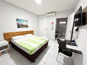 Quarto de hotel com cama, escrivaninha, cadeira, televisão e ar condicionado.