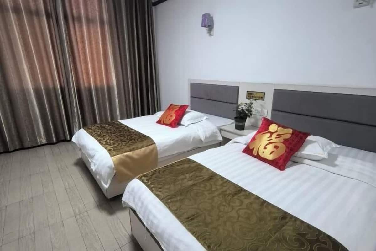 A hotel room with two beds, each with a white and gold bedspread, and red pillows with gold text. There is a nightstand with a vase of flowers and a wall-mounted light.