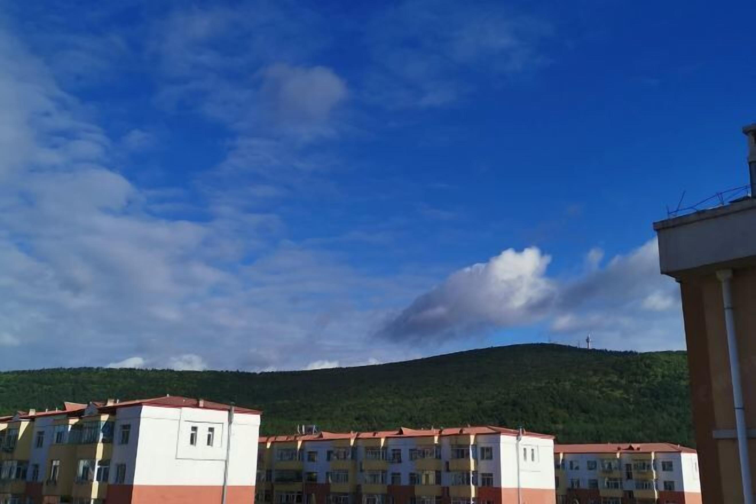 A multi-story building with a red roof, likely a hotel or residential complex, with a view of a green hill in the background.