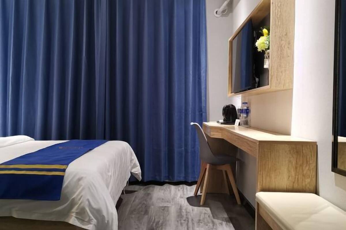 A hotel room with a bed, a desk with a chair, a mirror, and a window with curtains.