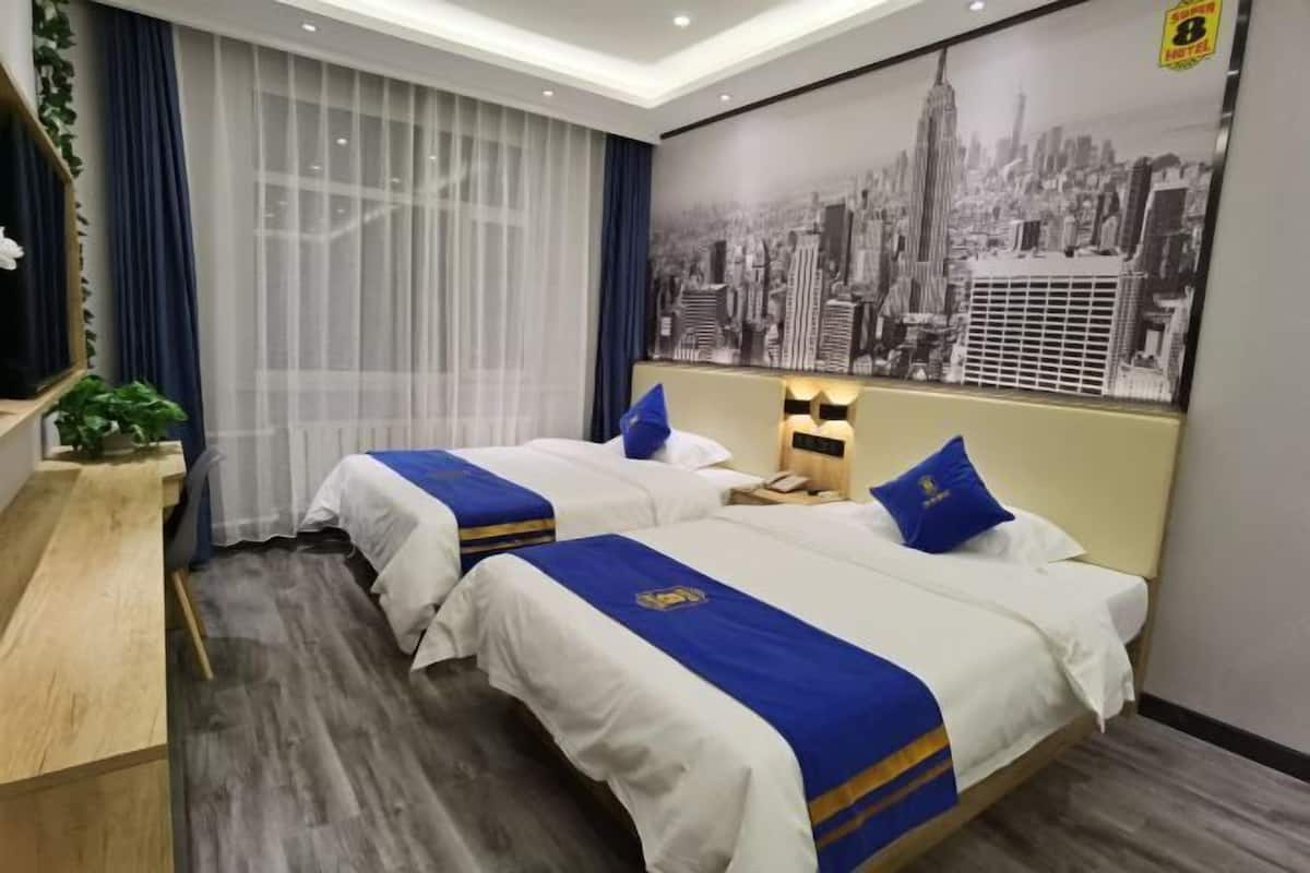 A hotel room with two beds, a desk, a large cityscape poster, and a window with sheer curtains.
