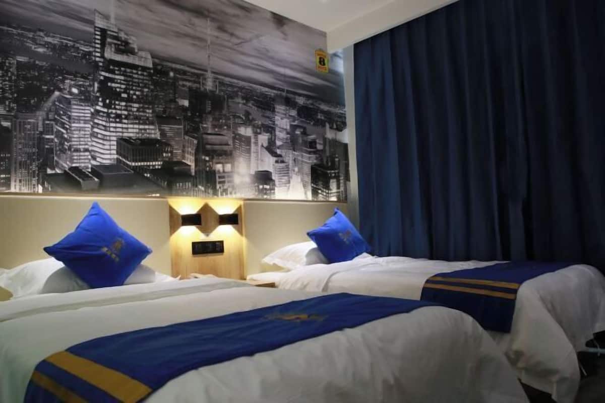A hotel room with two beds, a cityscape mural, a blue curtain, and a wall-mounted light fixture.