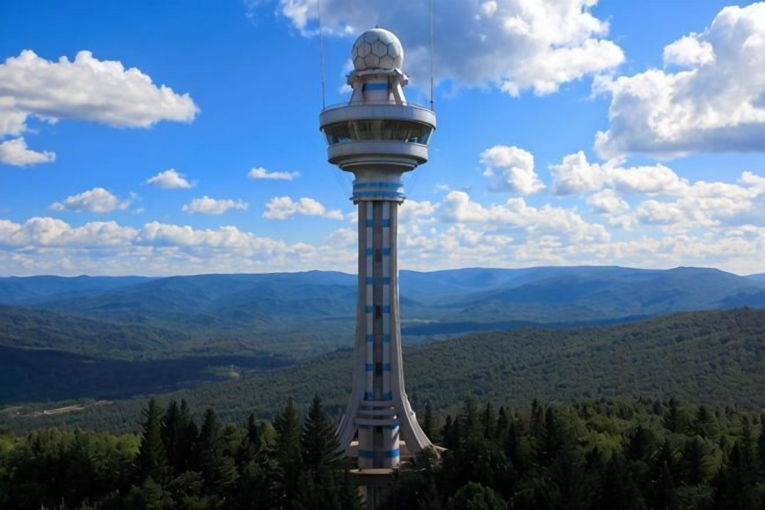 A tower with a spherical top, surrounded by forest and mountains.