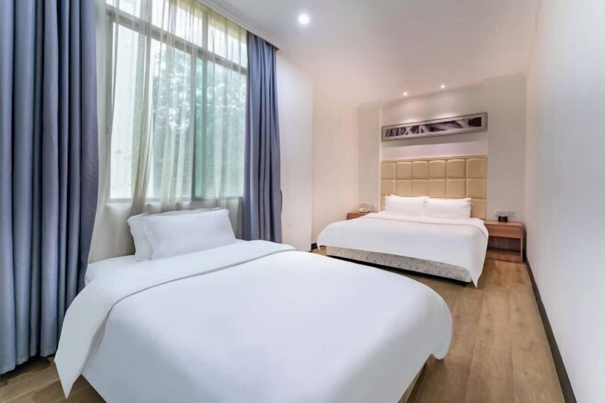 A hotel room with two beds, a large window with curtains, a wooden floor, and a wall-mounted air conditioning unit.