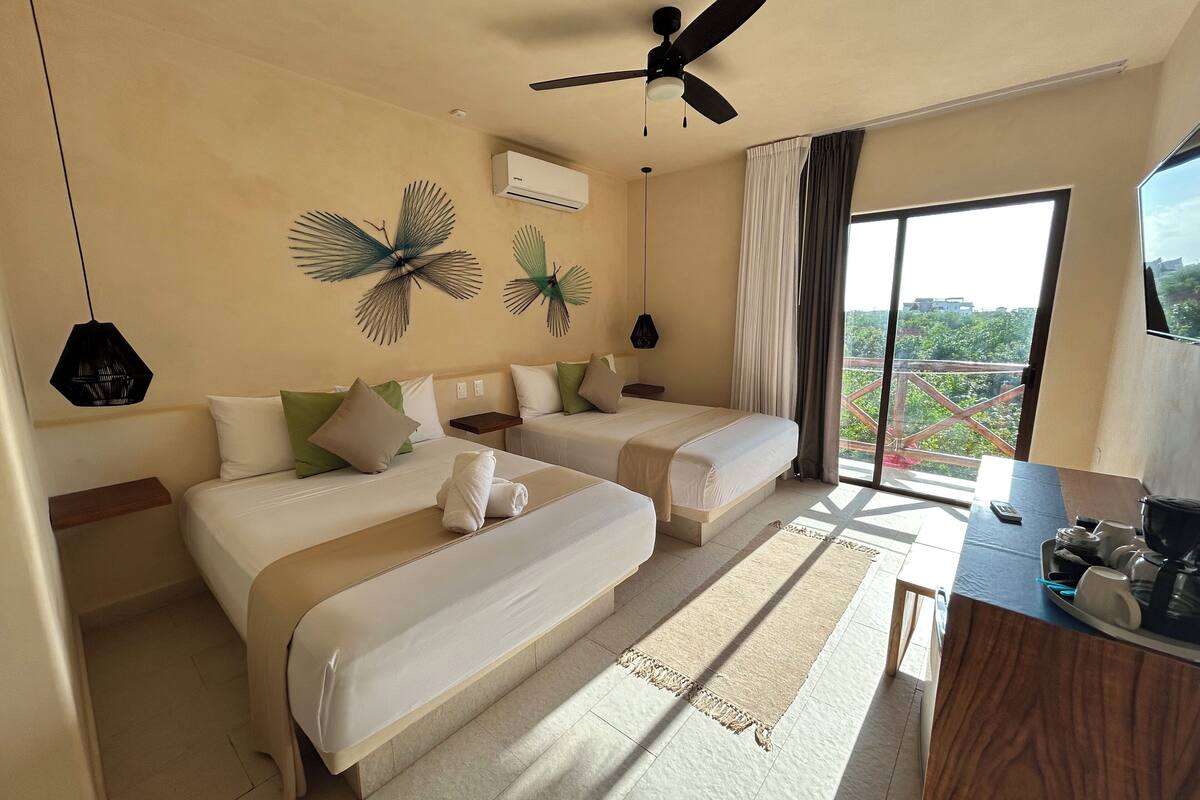 Quarto de hotel com duas camas, um ventilador de teto, aparelho de ar condicionado, uma janela grande com cortinas e vista para o verde lá fora.