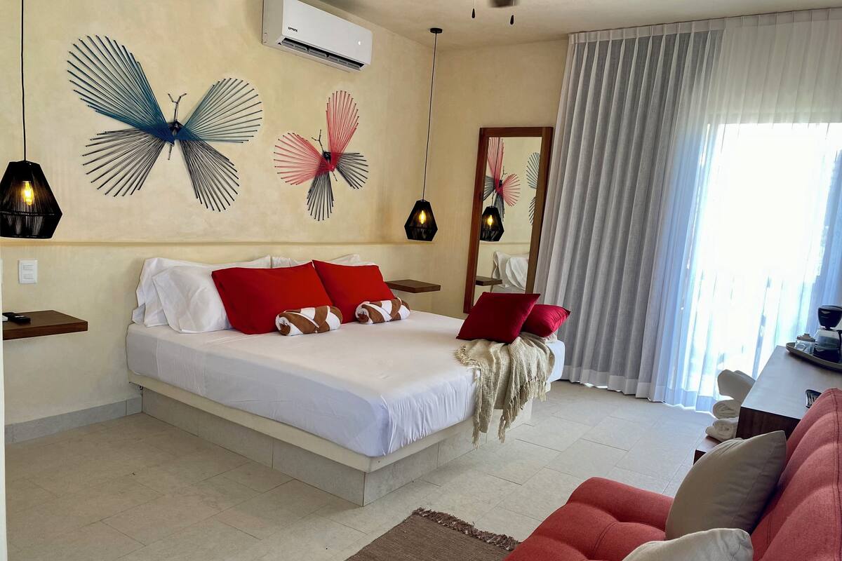 Quarto de hotel com uma cama grande, almofadas vermelhas e brancas, um sofá vermelho e arte decorativa na parede.