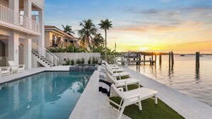 Un espace piscine moderne offrant une vue dégagée sur le coucher de soleil sur l’eau, un escalier menant aux niveaux supérieurs et une rangée de chaises longues blanches.