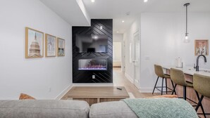 Un salon moderne avec une cheminée, un coin repas avec des tabourets de bar et une cuisine dotée d’un plan de travail blanc.