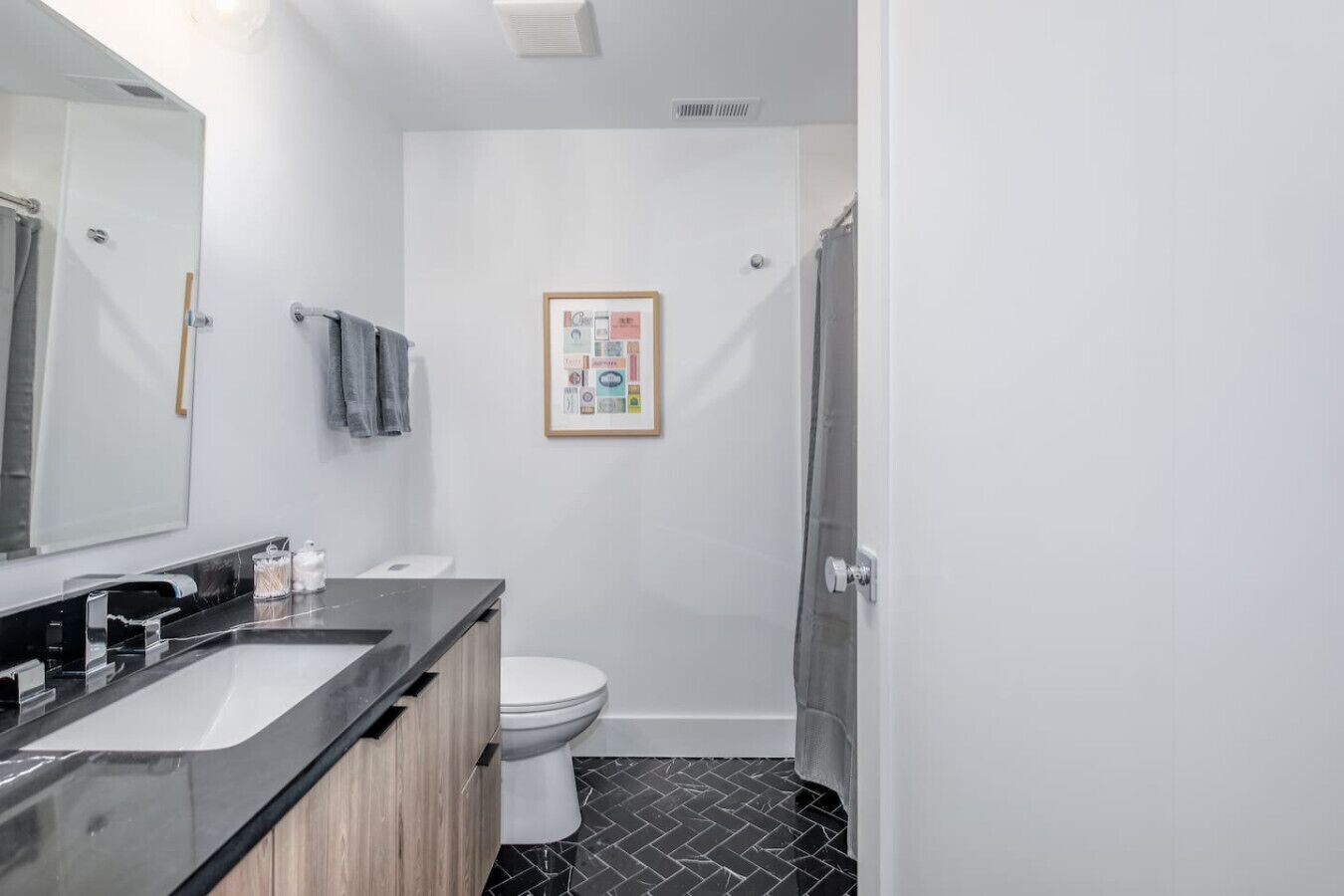 A modern bathroom with a black and white tiled floor, a white toilet, a black sink with a wooden cabinet, and a framed picture on the wall.