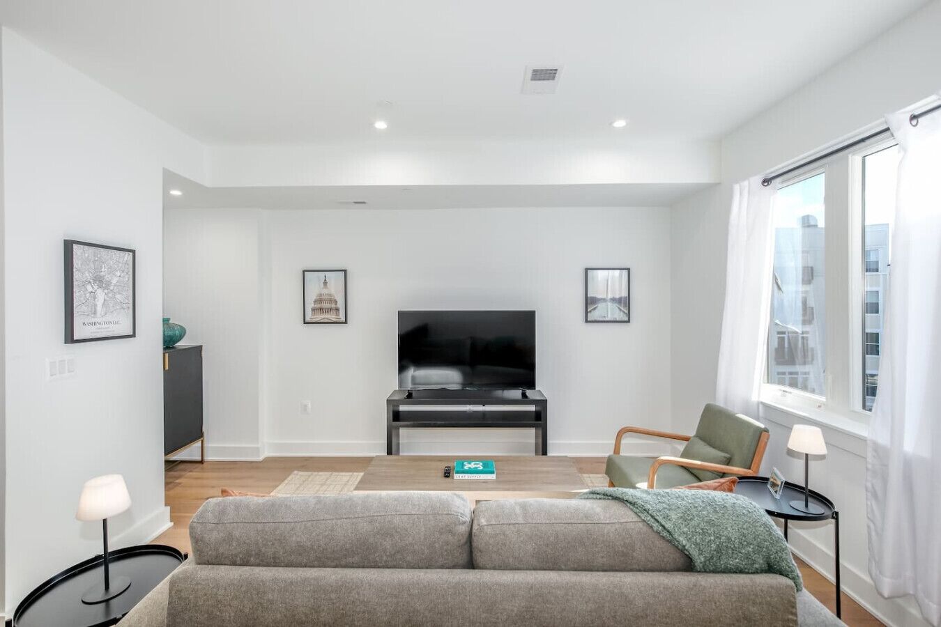 A modern living room with a flat-screen TV, grey sofa, and a large window with curtains.
