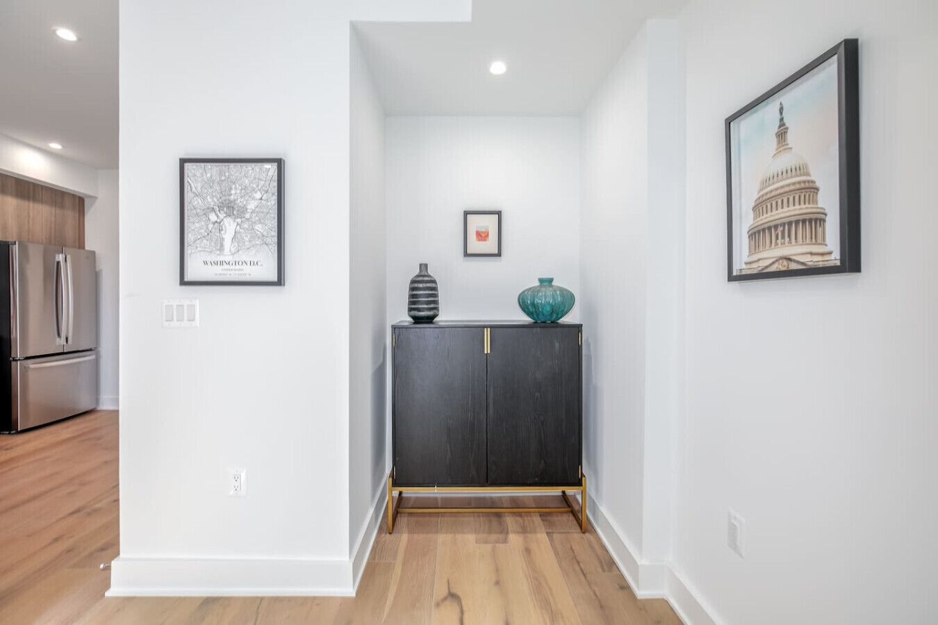 Un couloir avec une commode en bois sombre, un tableau encadré représentant un bâtiment et un tableau encadré représentant un arbre.