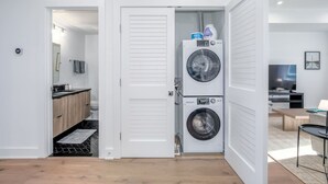 Une petite buanderie équipée de deux machines à laver à chargement frontal superposées, d’un petit évier et d’une porte donnant sur une salle de bain.