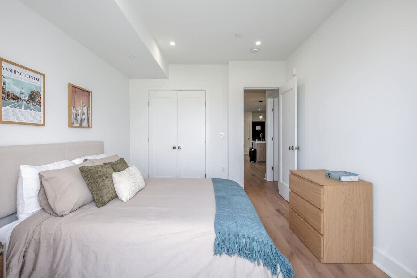 Quarto com cama, cómoda de madeira, porta e corredor que leva a um banheiro.