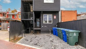 A two-story building with a black exterior, a visible staircase, and a row of blue and green recycling bins outside.